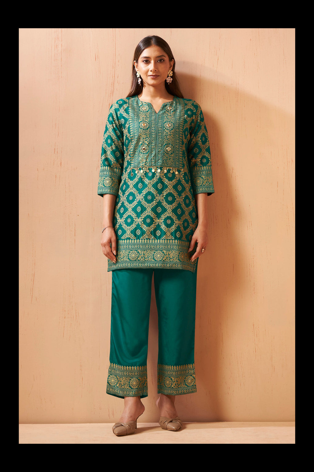 A woman modeling a traditional Indian outfit consisting of a teal green kurta with intricate gold and silver embroidery, paired with matching teal green pants, standing against a light wood-paneled wall.