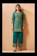 A woman modeling a traditional Indian outfit consisting of a teal green kurta with intricate gold and silver embroidery, paired with matching teal green pants, standing against a light wood-paneled wall.