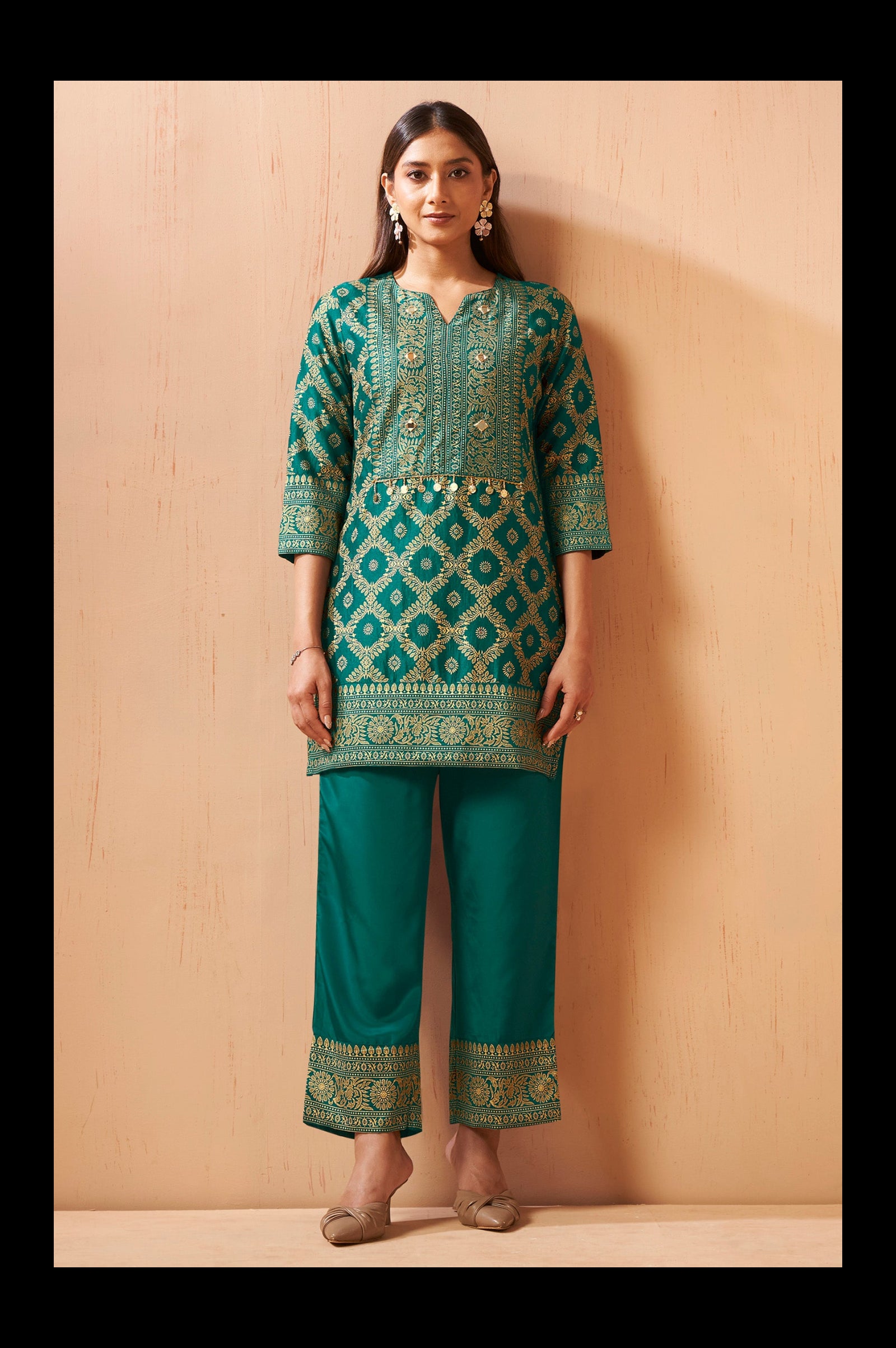 A woman modeling a traditional Indian outfit consisting of a teal green kurta with intricate gold and silver embroidery, paired with matching teal green pants, standing against a light wood-paneled wall.