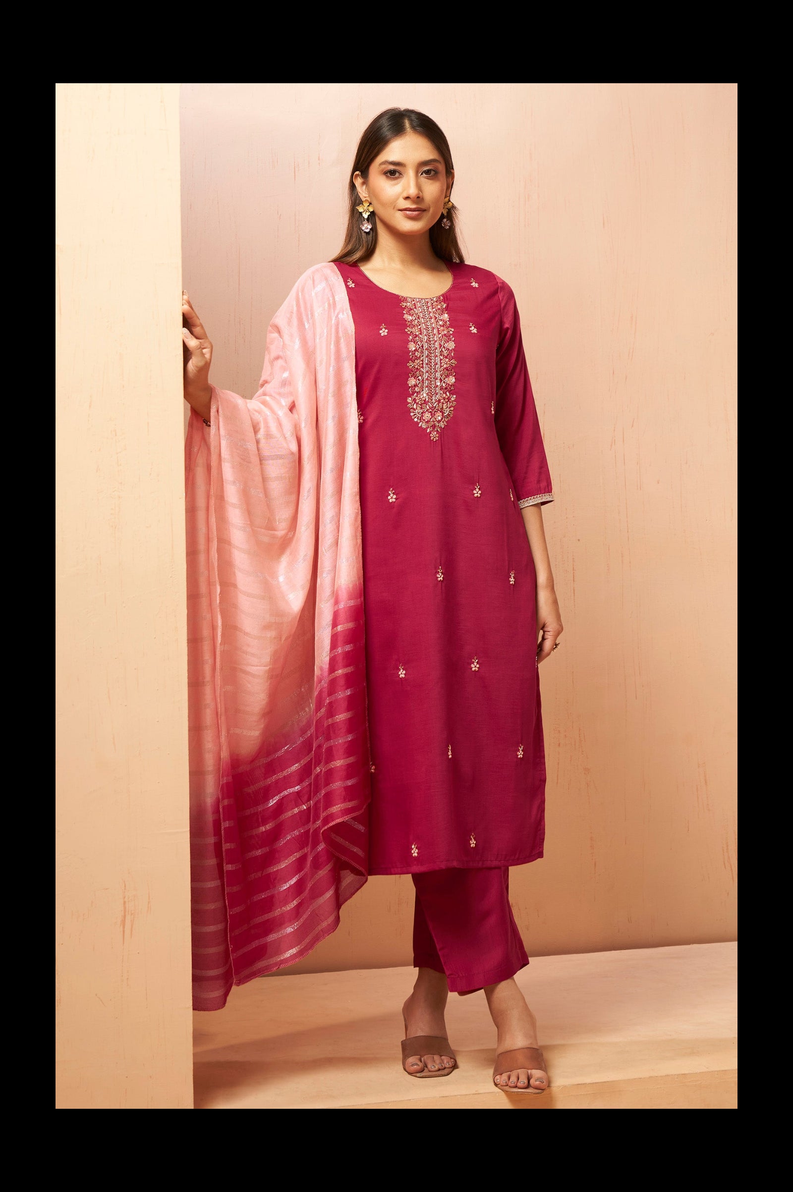 A woman models a traditional Indian salwar kameez suit set in a studio setting. The set includes a deep magenta kurta with gold embroidery on the neckline, matching trousers, and a light pink to magenta gradient striped dupatta.