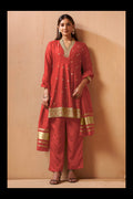 A woman modeling a red Indian ethnic suit set with gold embroidery, consisting of a kurta, pants, and a dupatta, standing against a beige textured wall.