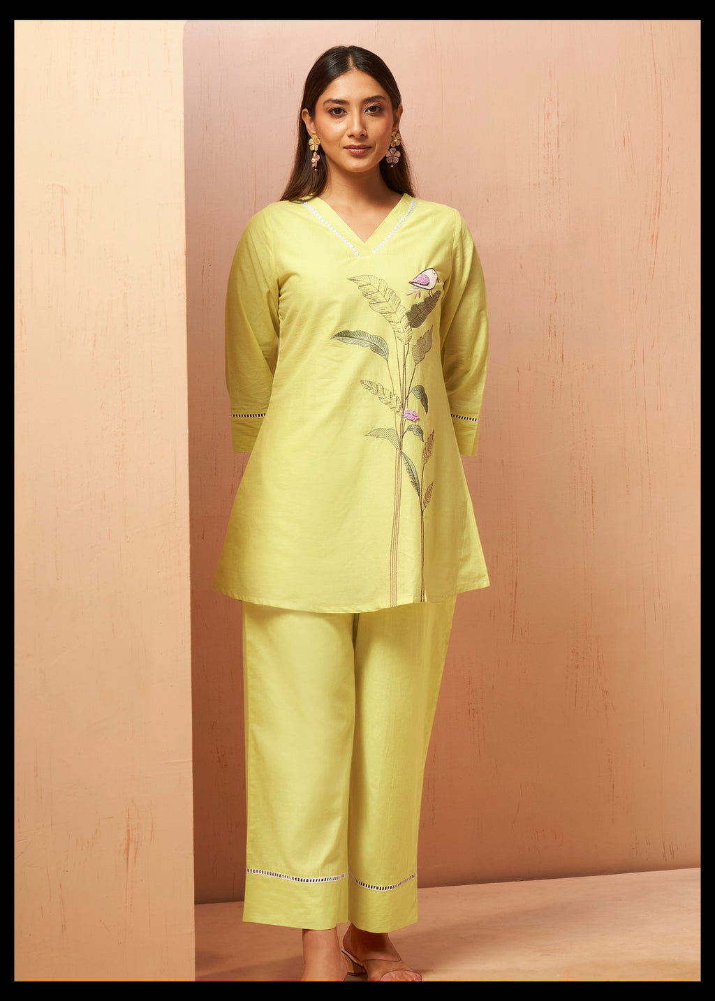 A woman modeling a light yellow-green kurta and trouser set with floral embroidery on the chest, standing against a beige textured backdrop.