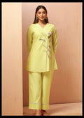 A woman modeling a light yellow-green kurta and trouser set with floral embroidery on the chest, standing against a beige textured backdrop.