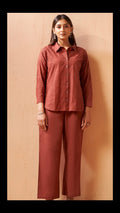 A woman stands against a light beige wooden wall wearing a matching rust-colored two-piece set consisting of a textured long-sleeve button-up shirt and wide-leg cropped pants.