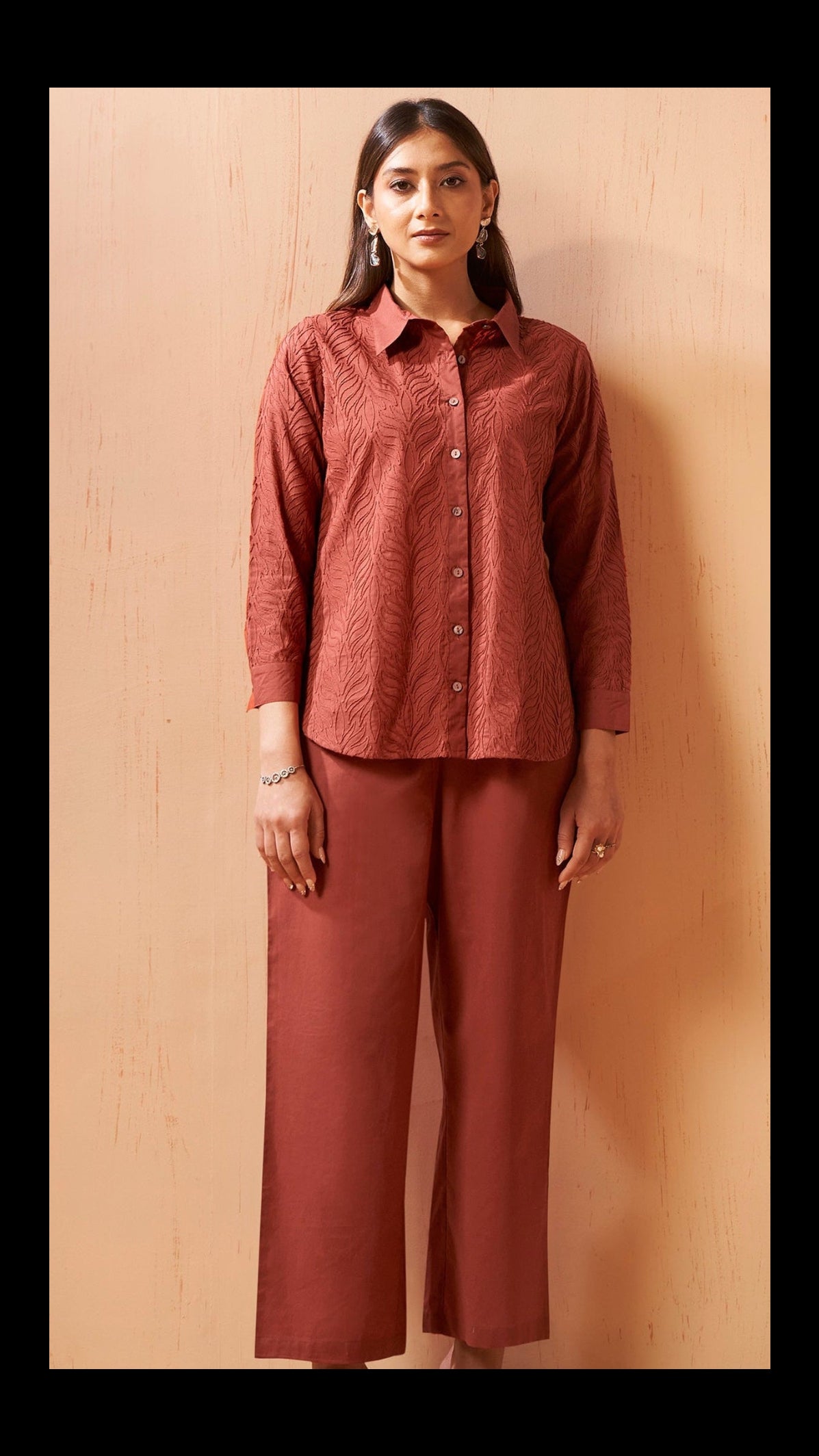 A woman stands against a light beige wooden wall wearing a matching rust-colored two-piece set consisting of a textured long-sleeve button-up shirt and wide-leg cropped pants.