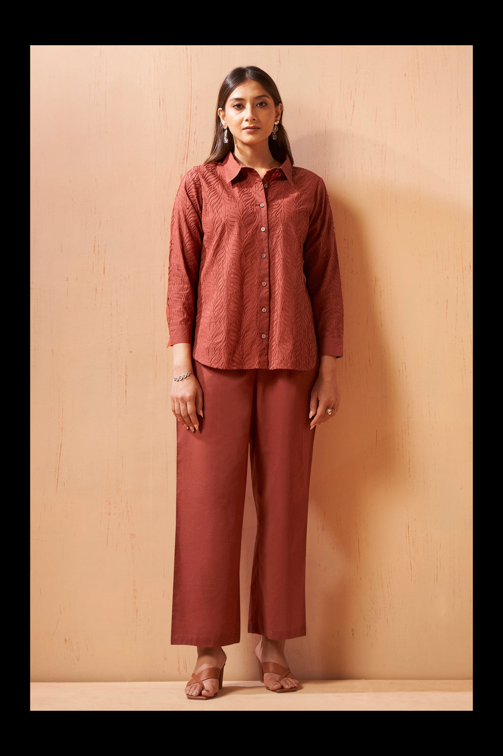 A woman stands against a light beige wooden wall wearing a matching rust-colored two-piece set consisting of a textured long-sleeve button-up shirt and wide-leg cropped pants.