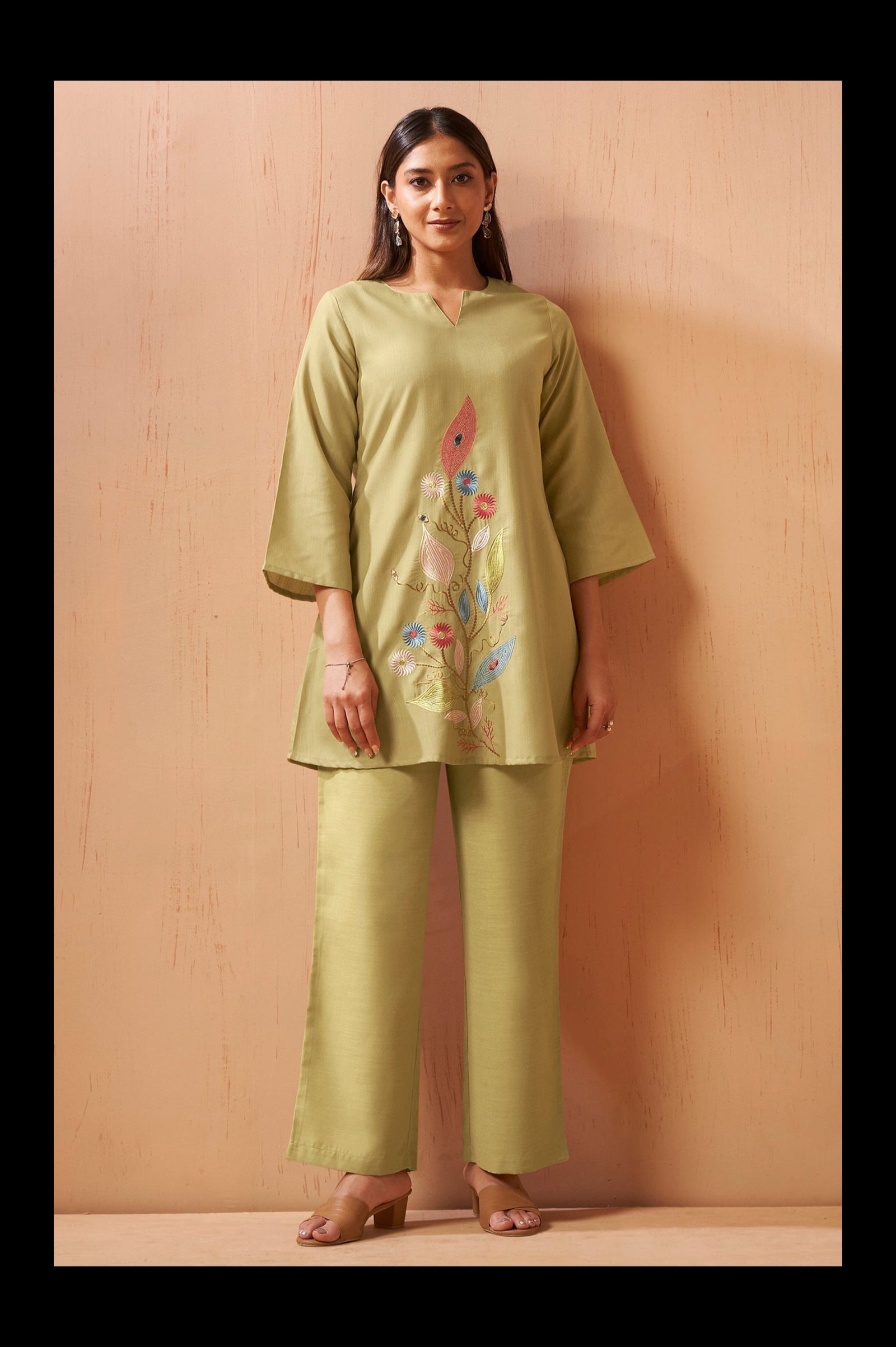 A woman modeling a sage green two-piece kurta and trouser set with colorful floral embroidery on the kurta, standing against a light wood-paneled wall.