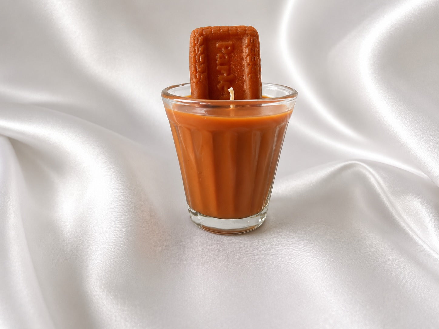 A small clear glass filled with orange-brown liquid, with a rectangular brown textured stick or bar standing upright inside the glass, set against draped white satin fabric.