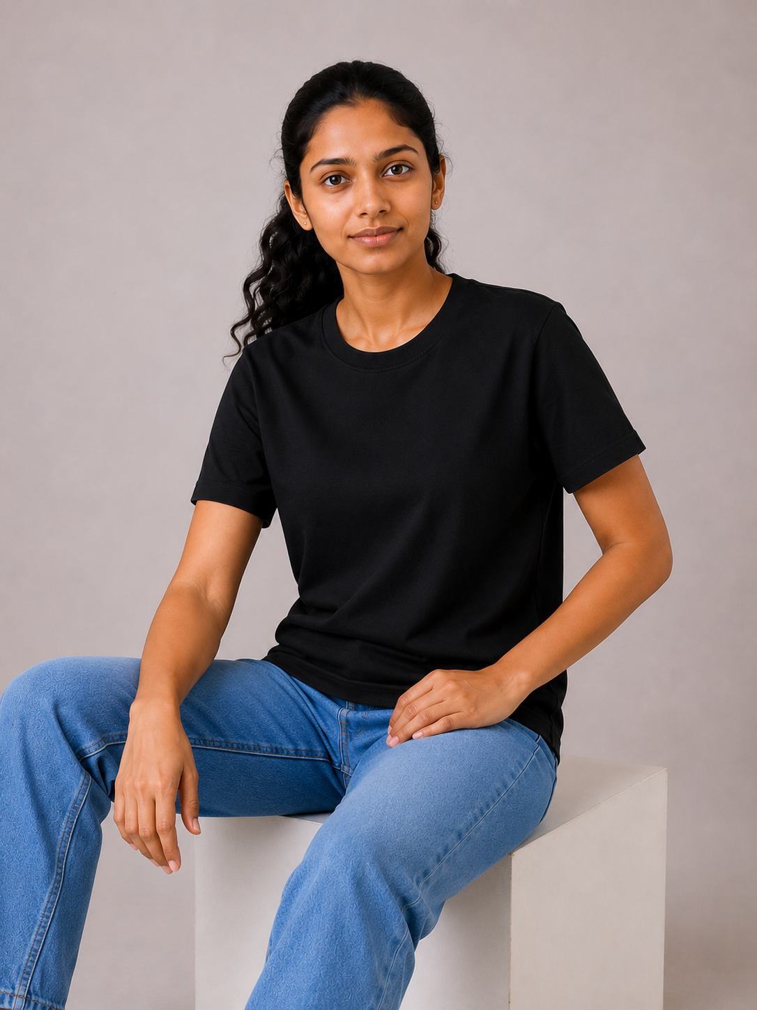 Woman wearing a black t-shirt and blue jeans sitting on a white block against a gray background