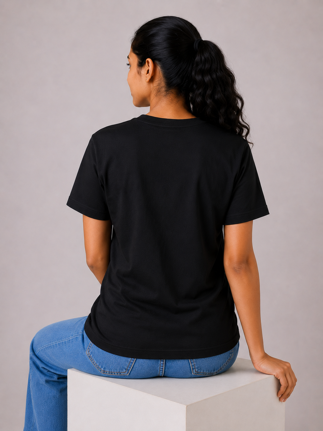Person wearing a black t-shirt and blue jeans sitting on a white cube against a gray background