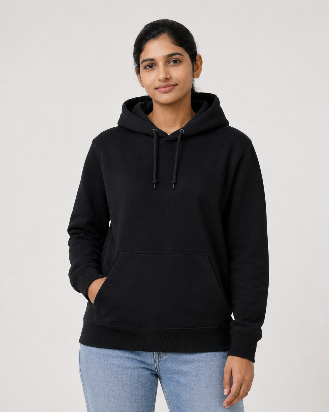 A woman wearing a black pullover hoodie with drawstrings and a front kangaroo pocket, paired with light blue jeans, standing against a plain light gray studio background.