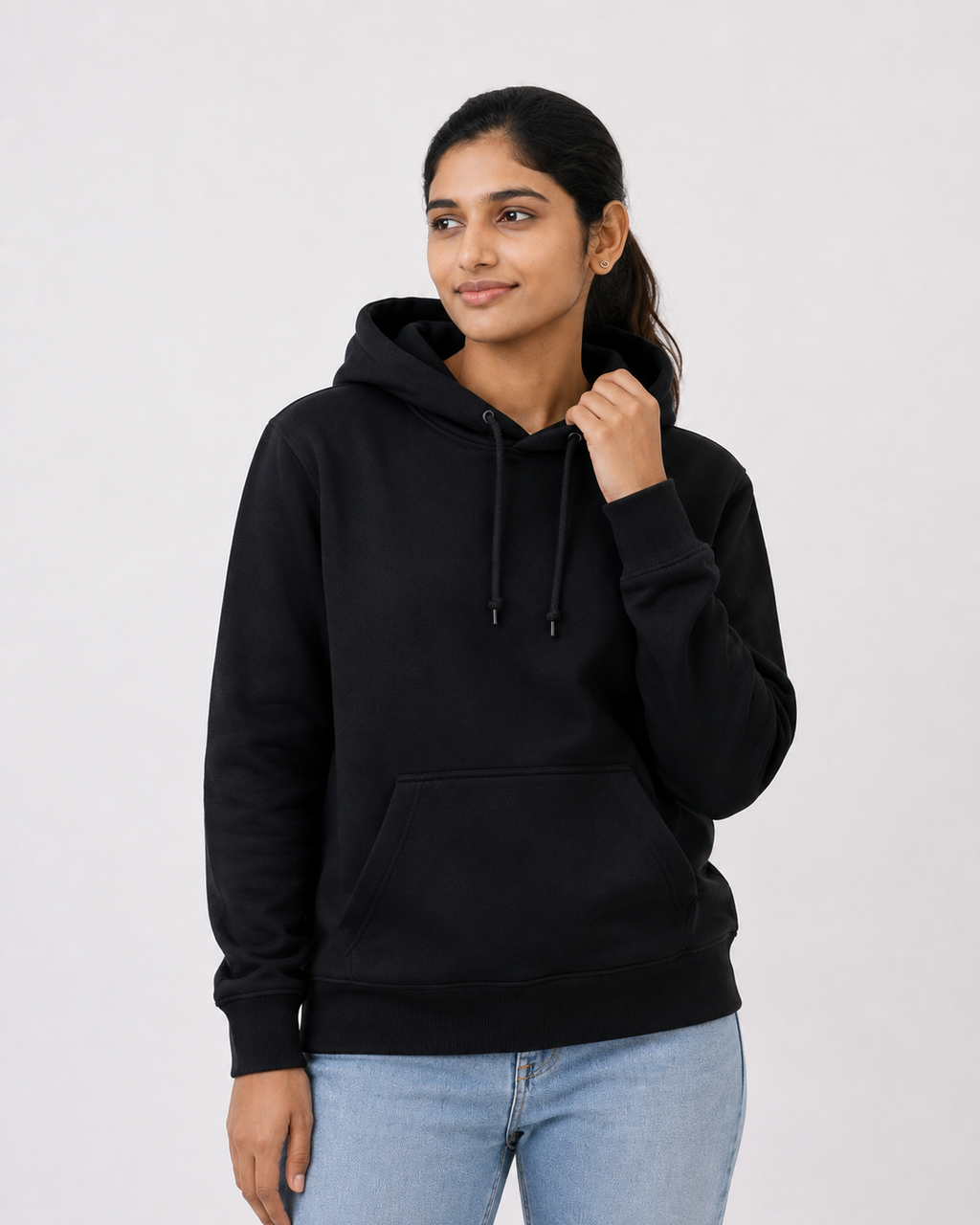 A woman wearing a black pullover hoodie with a front kangaroo pocket and black drawstrings, paired with light blue jeans, photographed against a plain light gray studio background.