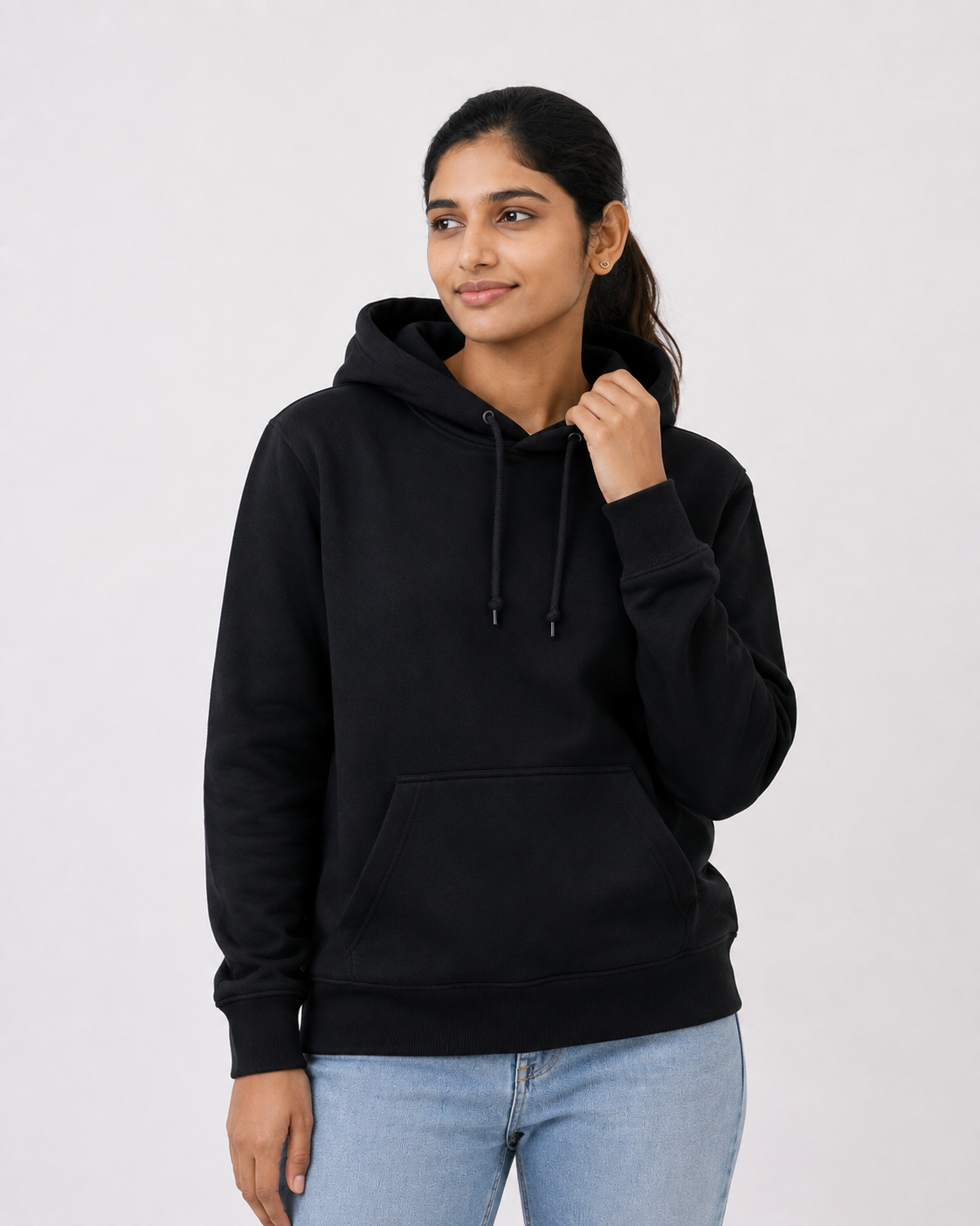 A woman wearing a black pullover hoodie with a front kangaroo pocket and black drawstrings, paired with light blue jeans, photographed against a plain light gray studio background.