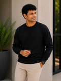 Man wearing a black sweater and light-colored pants standing indoors with a plant in the background