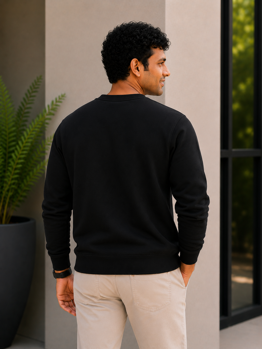 Man wearing a black sweatshirt and light-colored pants standing against a neutral wall with a plant in the background.