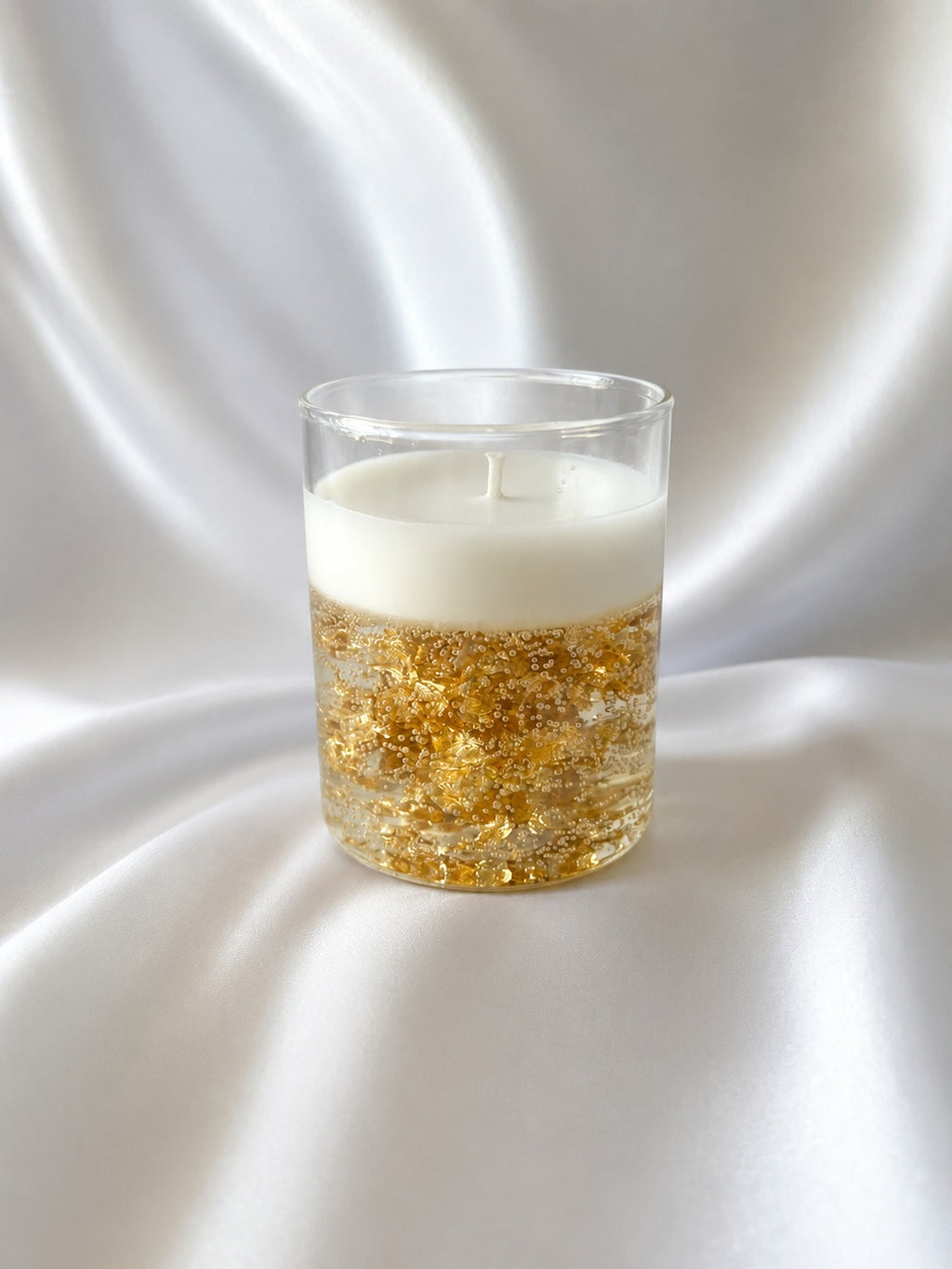A clear glass candle holder with a white wax top and a bottom filled with shimmering gold foil flakes, set against a draped white satin background.