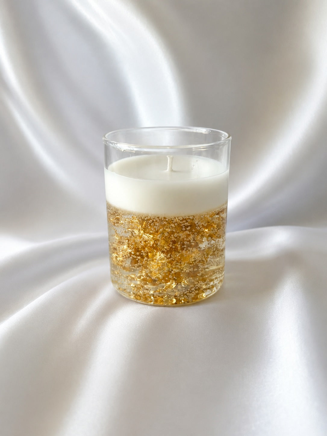 A clear glass candle holder with a white wax top and a bottom filled with shimmering gold foil flakes, set against a draped white satin background.