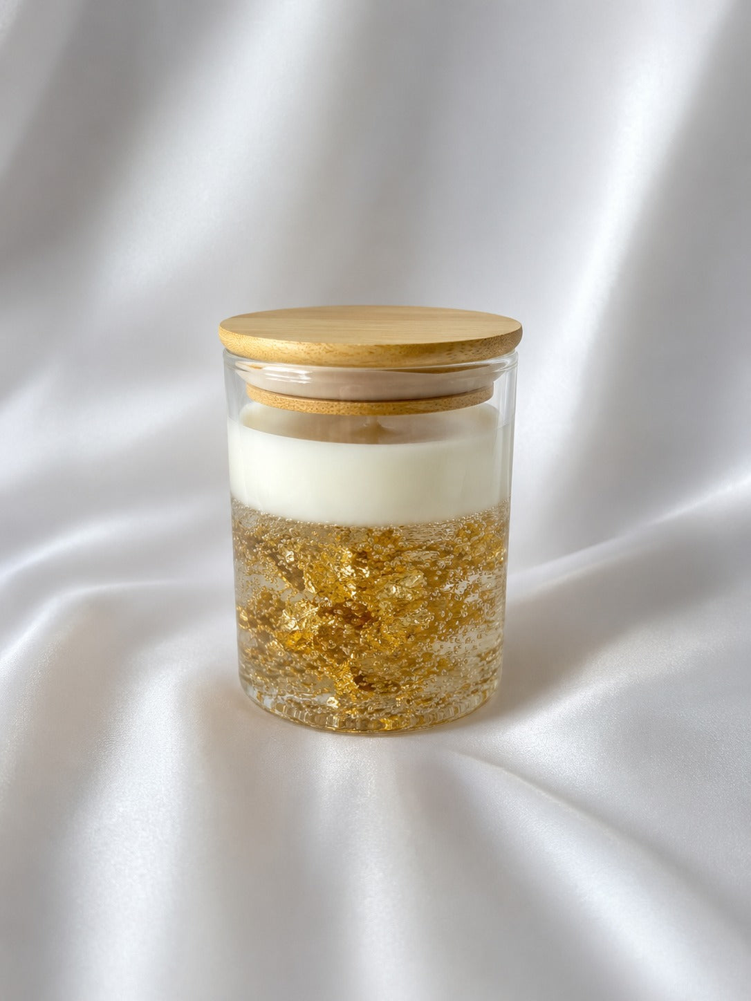 A clear glass jar candle with a light wood lid, featuring a white wax top and a bottom filled with shimmering gold leaf flakes, set against a draped white satin background.