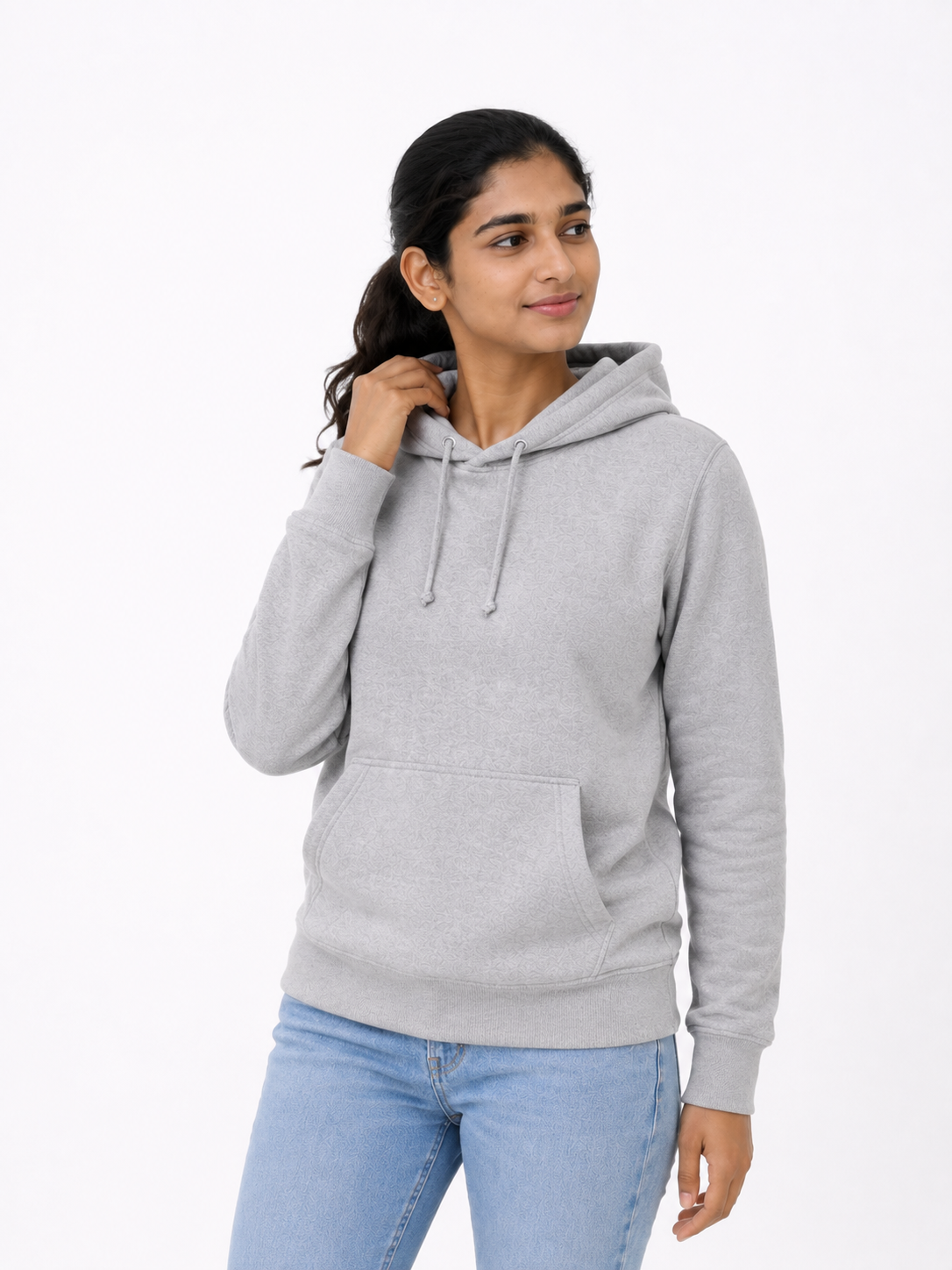 A woman wearing a light gray heather pullover hoodie with drawstrings and a front kangaroo pocket, paired with light blue jeans, against a plain white studio background.