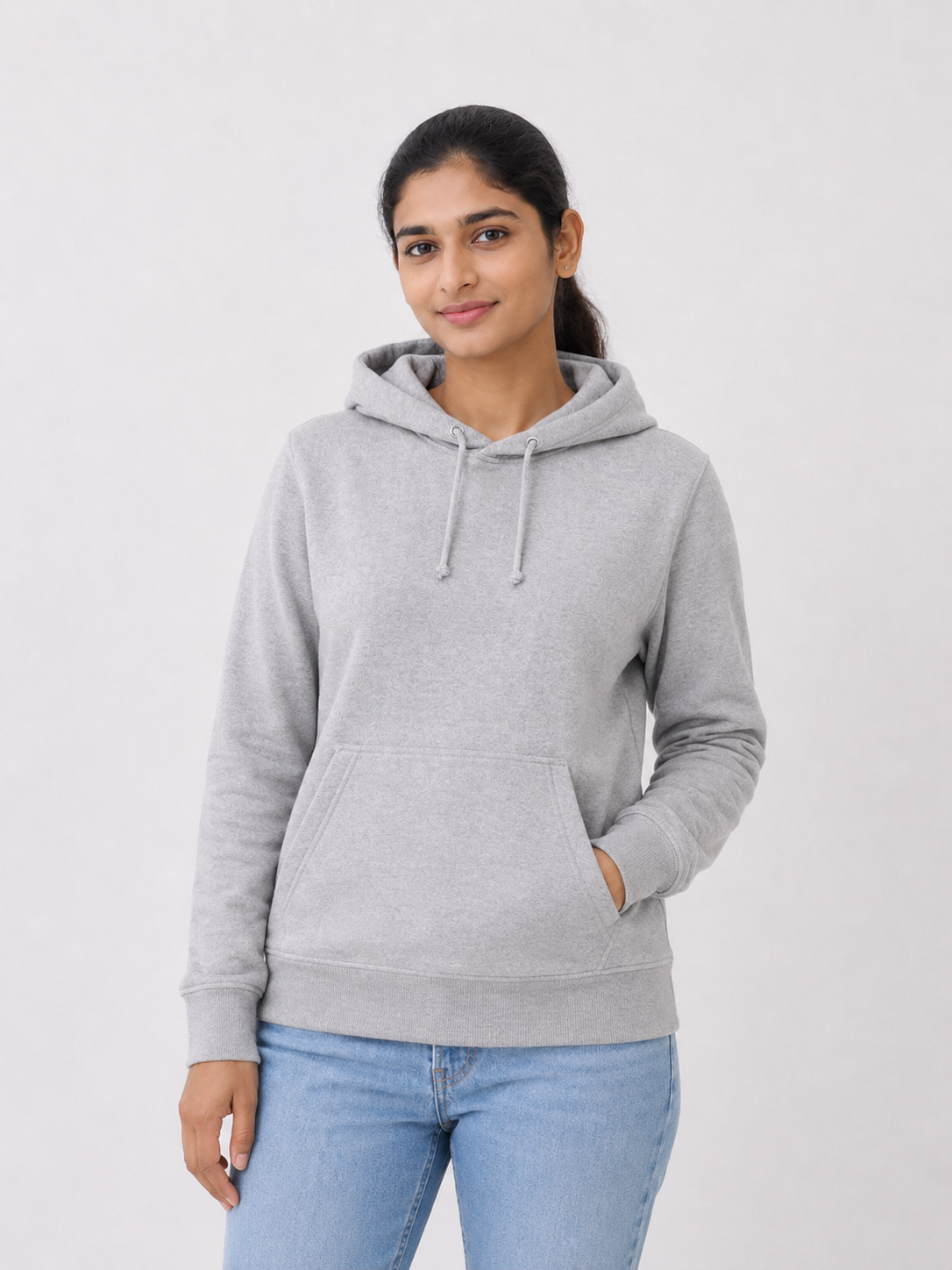 A woman wearing a light gray heather pullover hoodie with drawstrings and a front kangaroo pocket, paired with light blue jeans, standing against a plain light gray studio background.