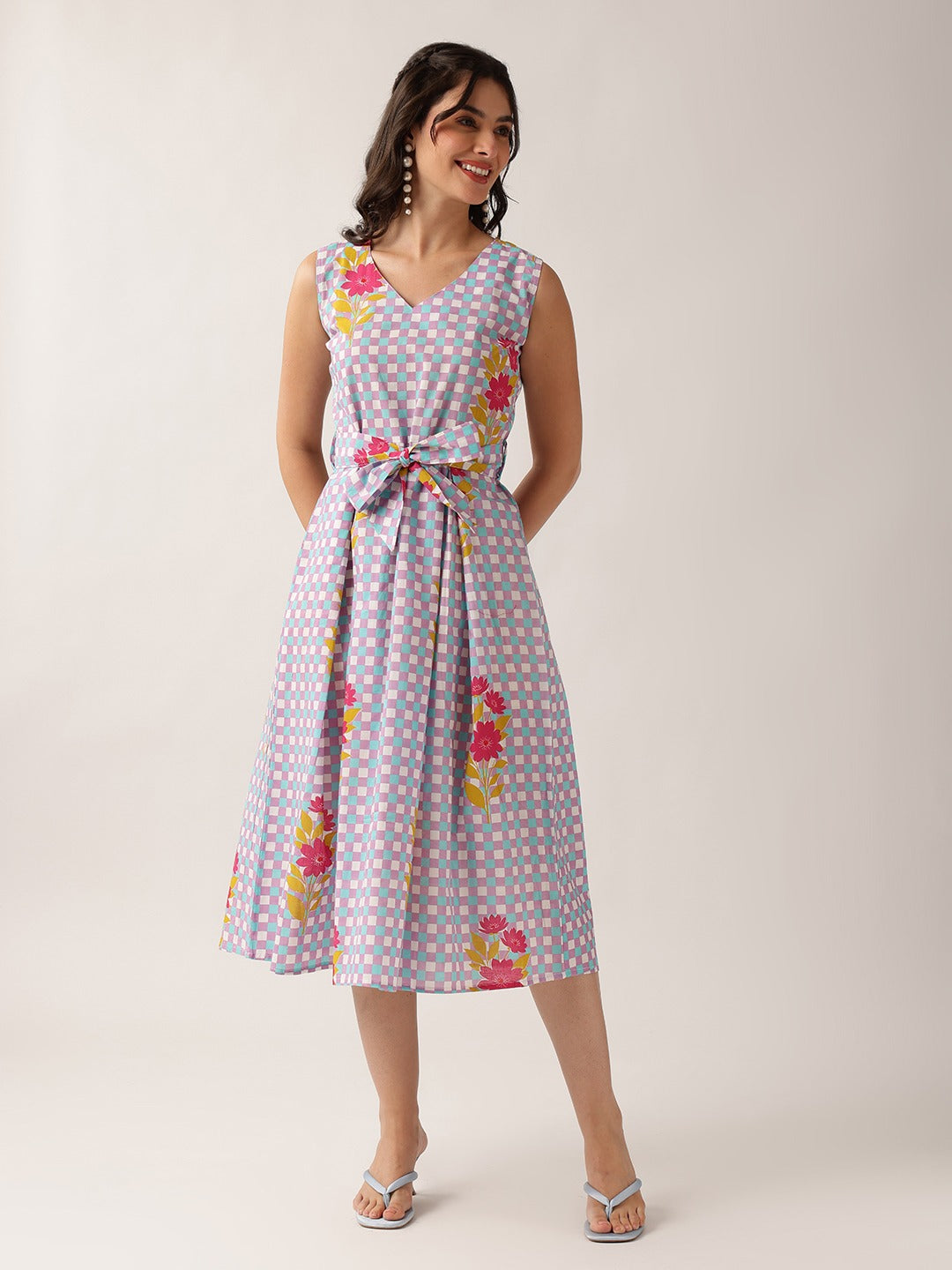 A woman modeling a sleeveless midi dress with a light blue and white gingham pattern and pink floral prints, featuring a tie waist and V-neckline, set against a plain light gray studio background.