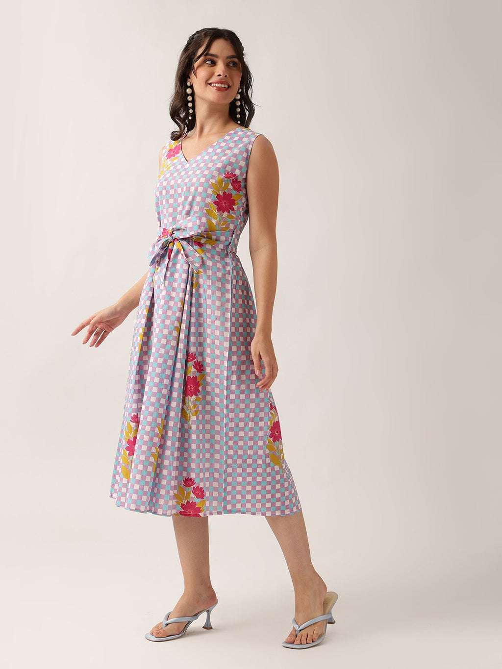 A woman modeling a sleeveless midi dress with a light lavender and pink gingham pattern and colorful floral prints, paired with silver heeled sandals against a plain light beige studio background.