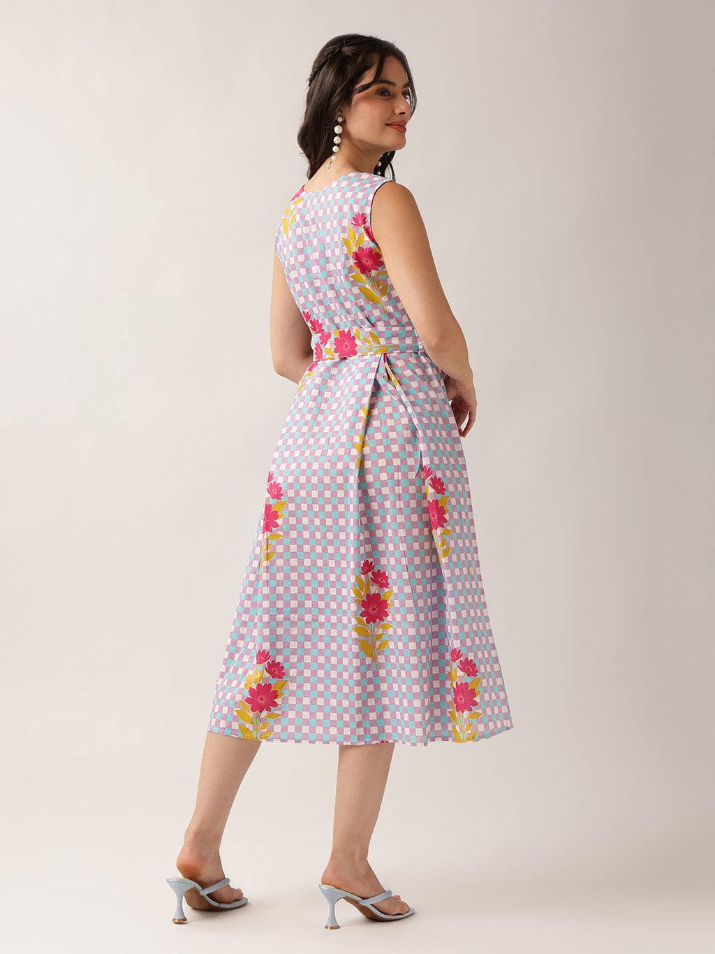 A woman modeling a sleeveless midi dress with a pastel blue and pink gingham pattern, yellow floral accents, and a matching tie belt at the waist, shown from a three-quarter back angle against a plain light beige studio background.