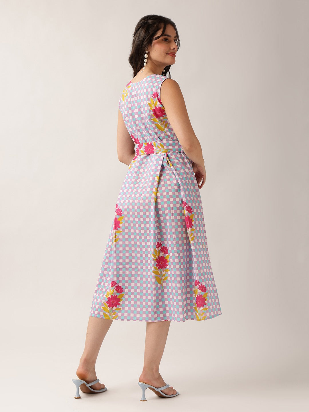 A woman modeling a sleeveless midi dress with a pastel blue and pink gingham pattern, yellow floral accents, and a matching tie belt at the waist, shown from a three-quarter back angle against a plain light beige studio background.
