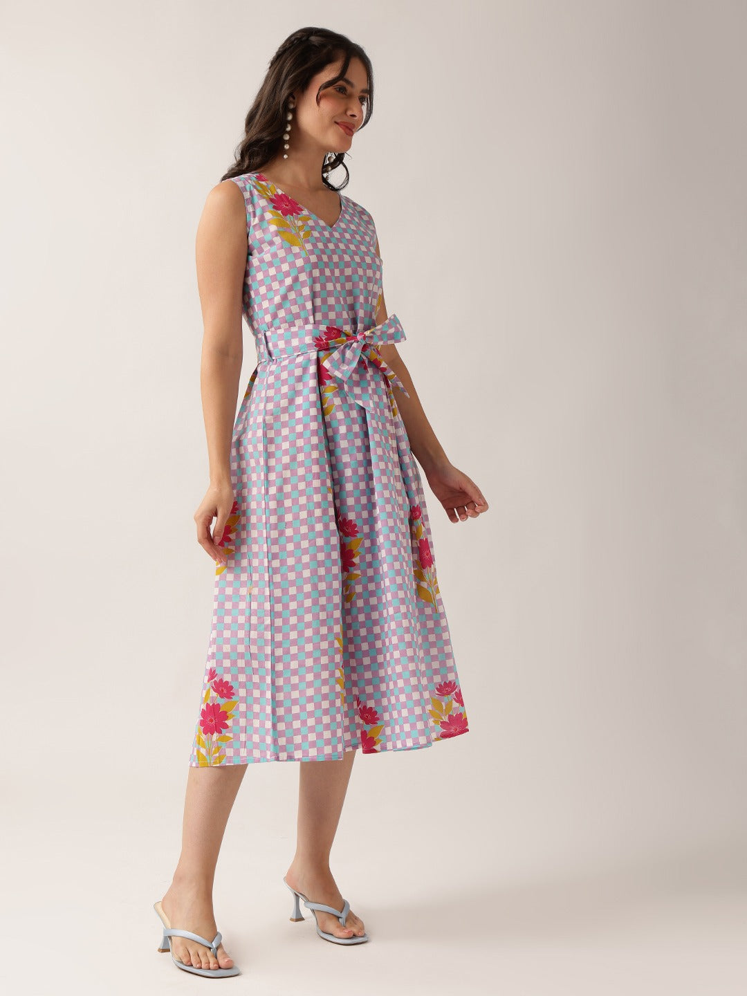 A woman modeling a sleeveless midi dress with a light purple and white gingham pattern, accented with pink and yellow floral prints and a matching tie belt at the waist, set against a plain light beige studio background.