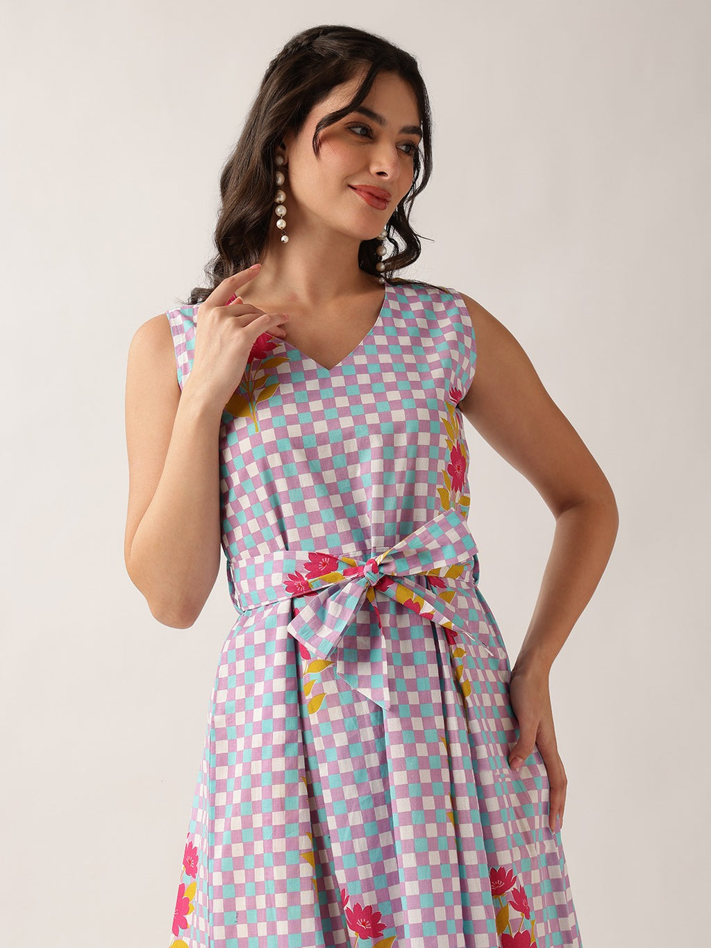 A woman modeling a sleeveless V-neck dress with a light blue, pink, and white checkered pattern and pink floral accents, tied at the waist with a matching bow, against a plain light gray background.