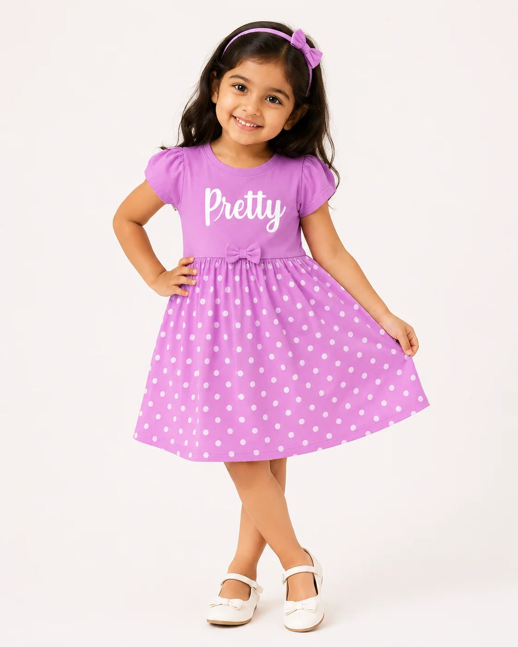 A young girl modeling a purple short-sleeve dress with white polka dots and the word 'Pretty' printed in white across the chest. She is wearing a matching purple bow headband and white shoes, standing against a plain white studio background.