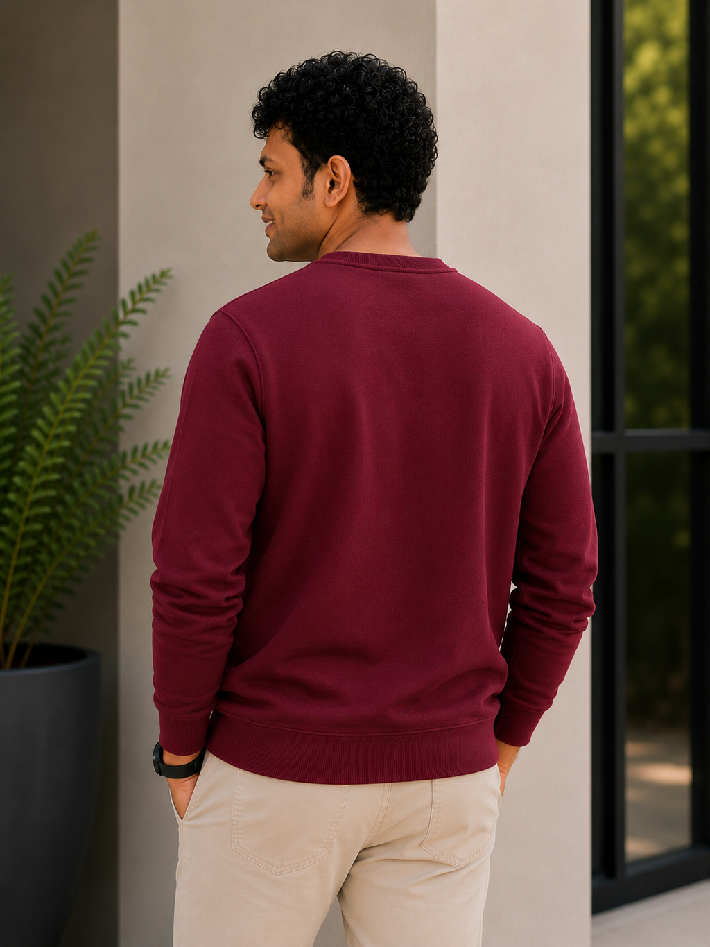 Man wearing a maroon sweater and beige pants standing against a neutral wall with a plant in the background.