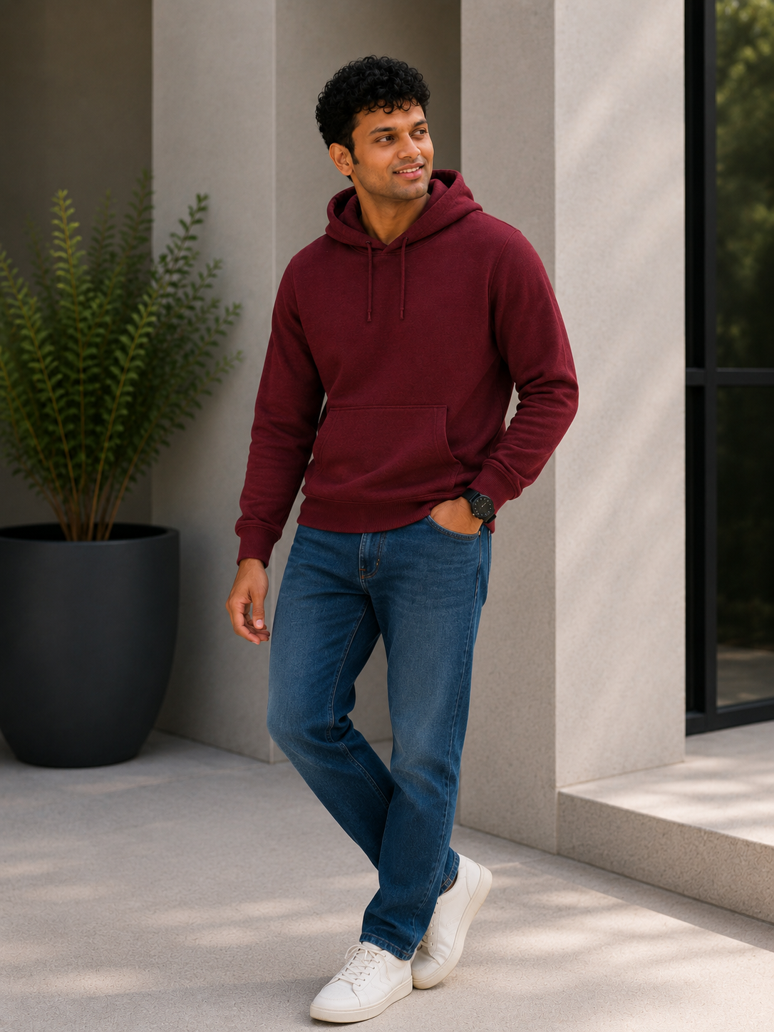 Man wearing a maroon hoodie and blue jeans standing outdoors near a building.