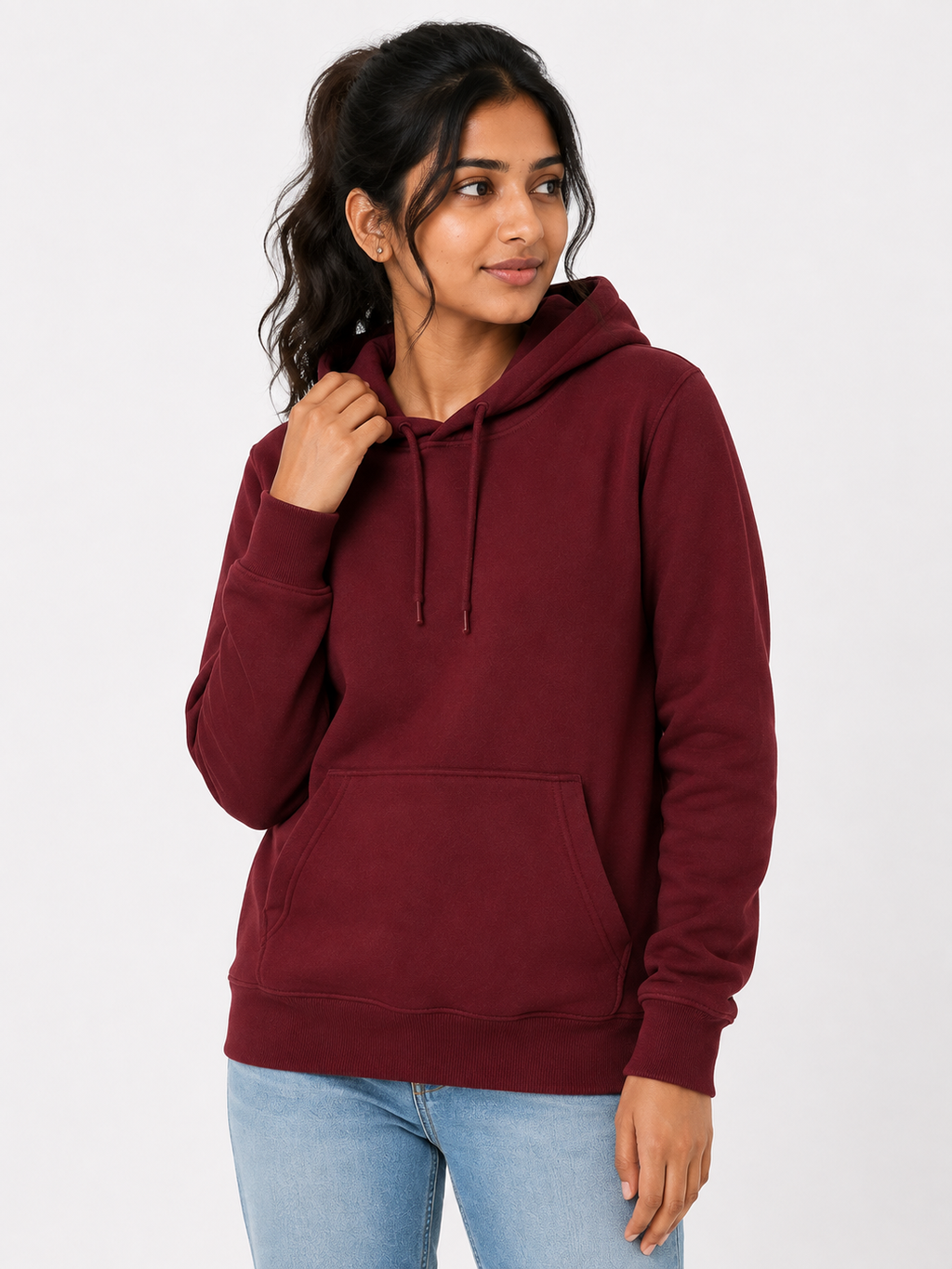 A woman wearing a maroon pullover hoodie with a front kangaroo pocket and matching drawstrings, paired with light blue jeans, against a plain light gray studio background.