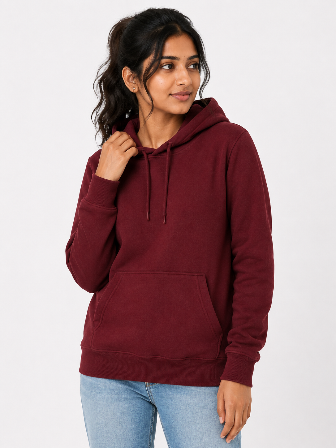 A woman wearing a maroon pullover hoodie with a front kangaroo pocket and matching drawstrings, paired with light blue jeans, against a plain light gray studio background.