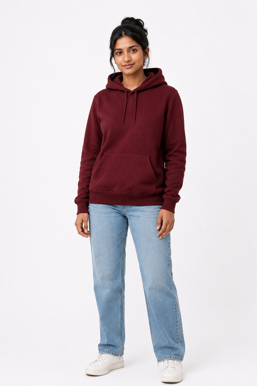 A woman modeling a burgundy pullover hoodie with light blue jeans and white sneakers, standing against a plain white studio background.