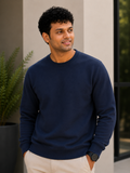 Man wearing a navy blue sweater standing outdoors with plants and a building in the background