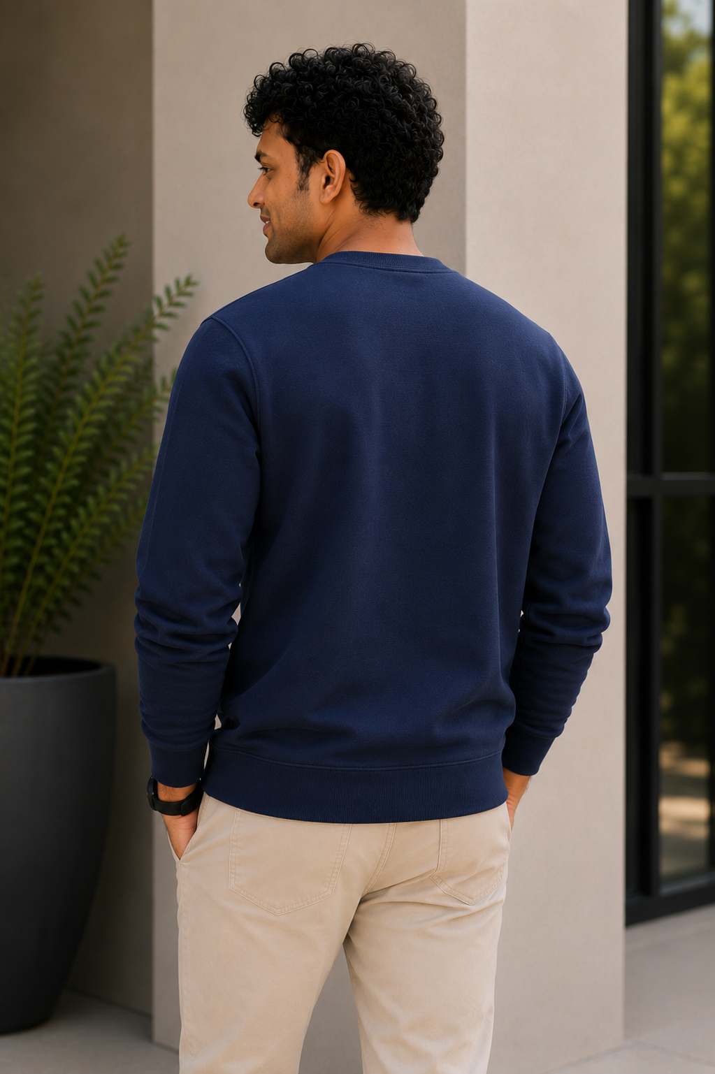 Man wearing a navy blue sweater and beige pants standing against a neutral wall with a plant in the background.