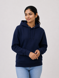 A woman wearing a navy blue pullover hoodie with a drawstring hood and kangaroo pocket, paired with light blue jeans, standing against a light gray studio background.
