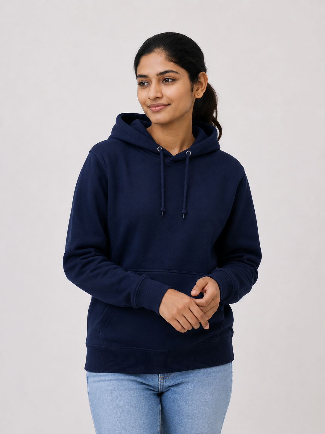 A woman wearing a navy blue pullover hoodie with a drawstring hood and kangaroo pocket, paired with light blue jeans, standing against a light gray studio background.