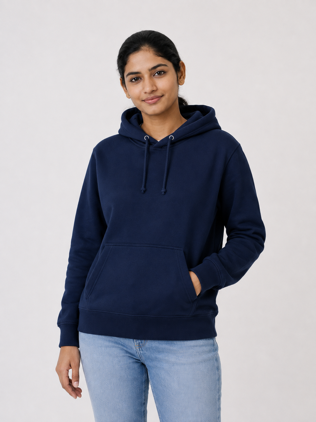 A woman wearing a navy blue pullover hoodie with drawstrings and a front kangaroo pocket, paired with light blue jeans, standing against a plain light gray studio background.
