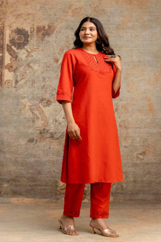 Woman wearing an orange kurta with embroidery against a textured wall.