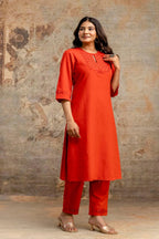 Woman wearing an orange kurta with embroidery against a textured wall.