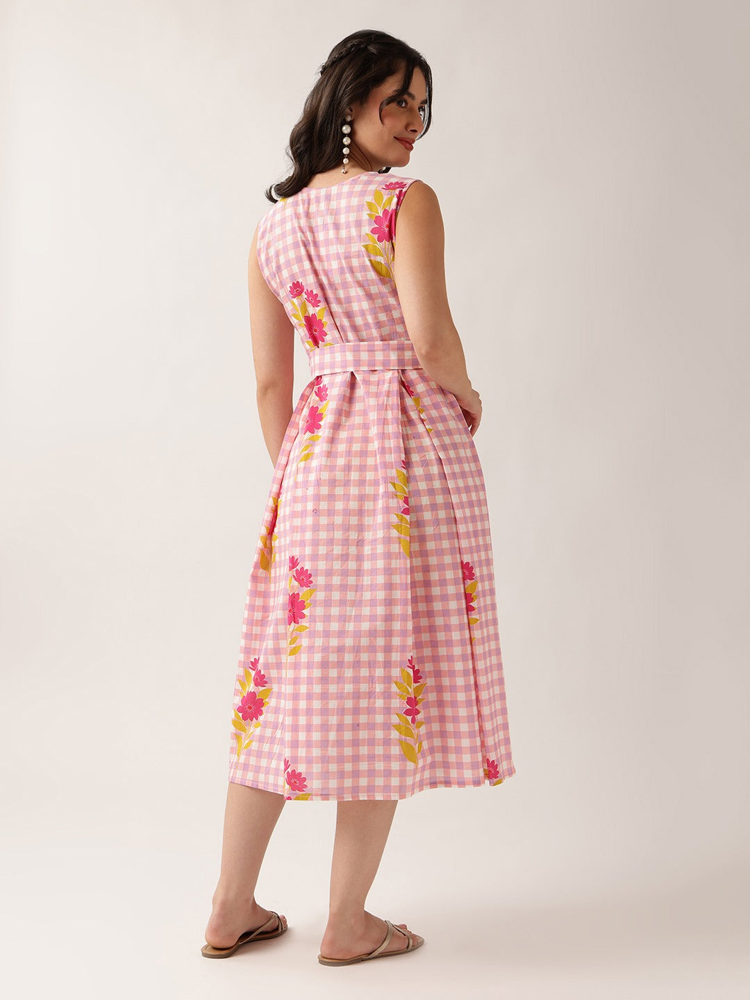 A woman modeling a sleeveless pink gingham midi dress with a waist tie and pink, yellow, and green floral embroidery, shown from a back-three-quarter view against a light beige studio background.