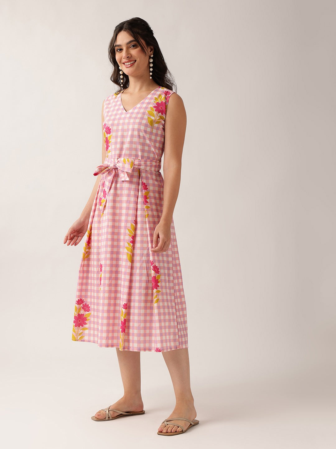 A woman modeling a sleeveless pink and white gingham midi dress with a matching waist tie and pink, yellow, and green floral accents, set against a plain light beige studio background.