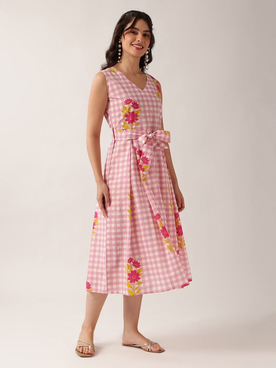 A woman models a sleeveless pink and white gingham midi dress with a matching tie belt and pink, yellow, and green floral accents, set against a plain light beige studio background.