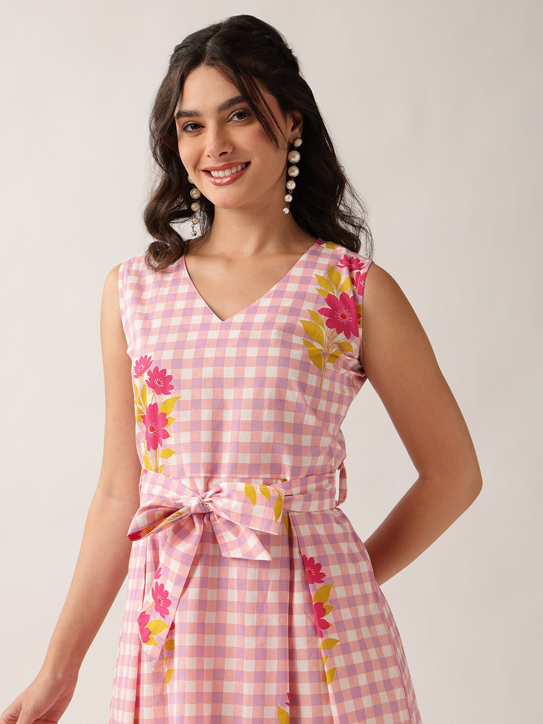 A woman wearing a sleeveless pink and white gingham dress with pink and yellow floral prints, featuring a matching tie belt at the waist, set against a plain light background.