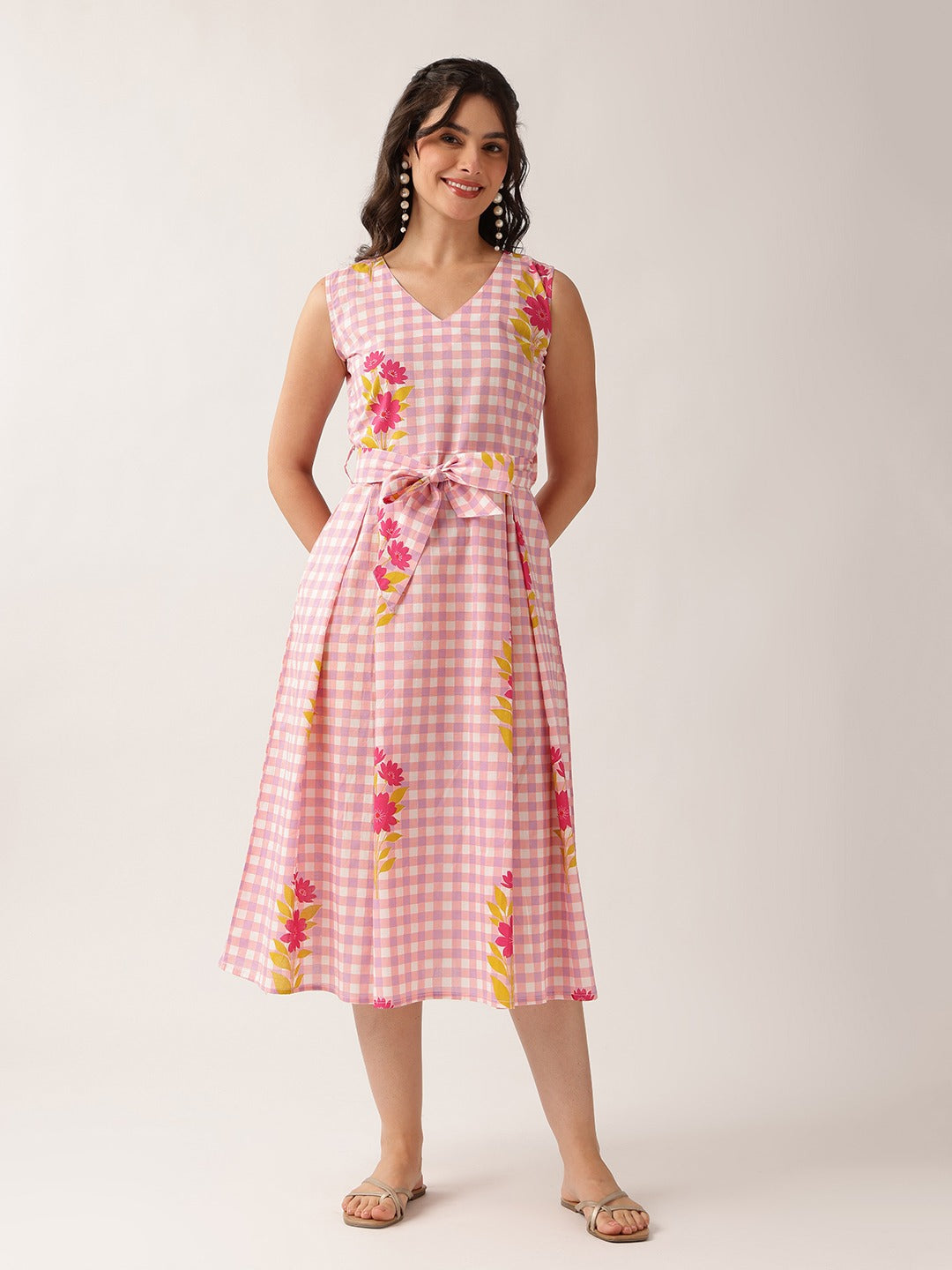 A woman modeling a sleeveless pink gingham midi dress with a tie waist and pink, yellow, and green floral print, set against a plain light beige studio background.