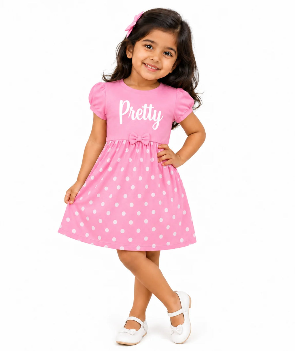 A young girl modeling a pink short-sleeve dress with white polka dots and the word 'Pretty' printed in white script across the chest. She is wearing white shoes and a pink hair clip, posing against a solid white background.
