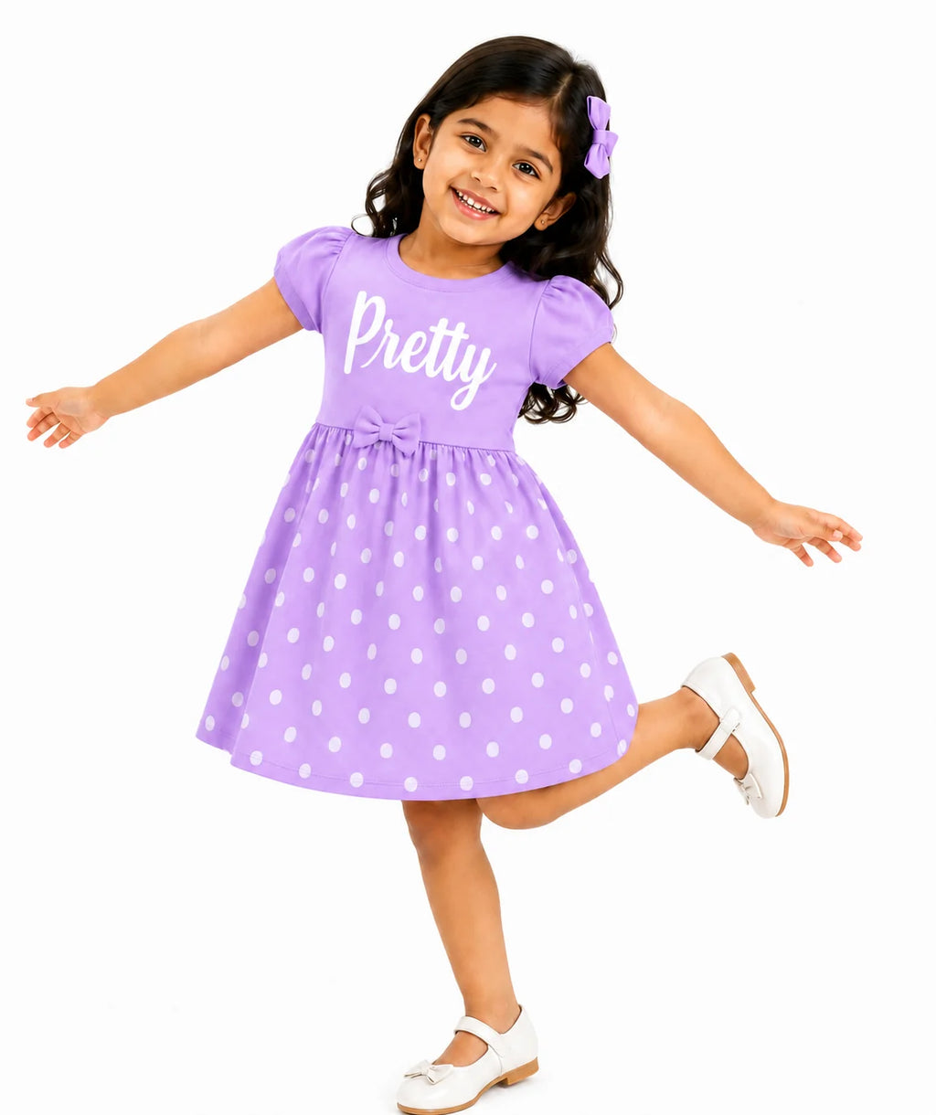 A young girl with dark hair wearing a purple short-sleeve dress with white polka dots and the word 'Pretty' printed on the chest, posing against a solid white background.